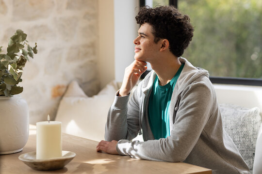 African male sitting at wood table dining nook, resting chin, gazing out window near candle pot