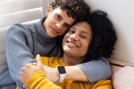 African American couple hugging on sofa at window with blinds, wearing knit sweaters and smartwatch