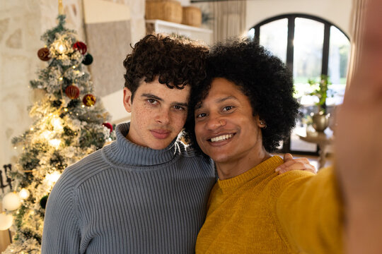 Diverse male couple wearing sweaters taking selfie with phone in living room near Christmas tree