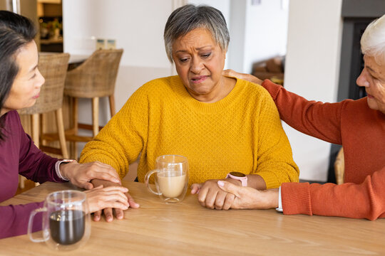 Diverse female friends supporting upset senior woman at kitchen table with mugs and wristwatch