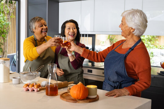 Diverse female friends wearing aprons clinking glasses and prepping dough in home kitchen with bowl