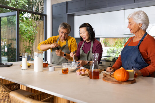 Diverse female friends whisking batter, prepping pumpkin on island, wearing aprons and watches