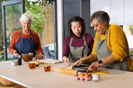 Diverse women wearing aprons working at kitchen island preparing pastry with tart pan, rolling pin