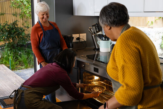 Diverse female friends wearing aprons, baking together in home kitchen, placing pie tin in oven