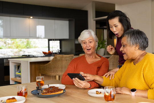 Diverse female friends leaning, viewing smartphone in modern kitchen with crumb cake and iced tea