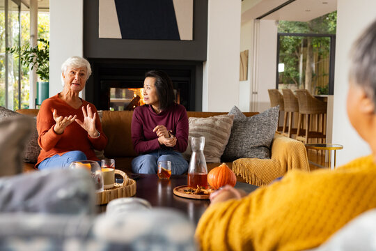 Diverse female friends sitting and chatting around wooden coffee table in living room, holding tea