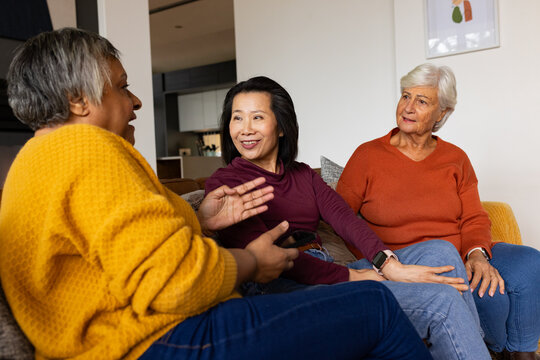 Diverse female friends chatting on couch in living room, holding smartphone and wearing jeans