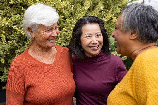 Diverse female friends with senior woman laughing and chatting near shrubbery wearing knit sweaters