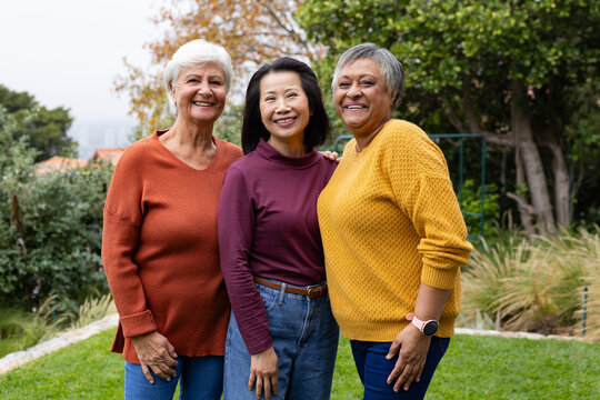 Diverse female friends standing in backyard lawn wearing knit sweaters, brown belt, smartwatch