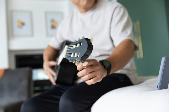 Senior Asian man holding black classical guitar, tuning strings, wearing white tee, phone nearby