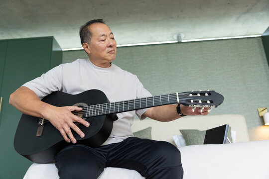 Senior Asian man sitting on bed edge holding and tuning black classical guitar with tablet nearby