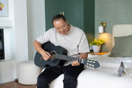 Senior Chinese man sitting on bed wearing beige tee playing black guitar with tablet nearby