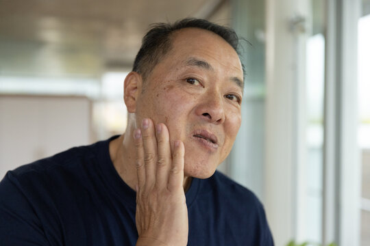 Senior Asian man wearing navy shirt raising right hand inspecting cheek in hall near window frames