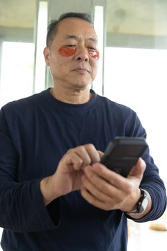 Senior Asian man standing holding phone in room in navy shirt wristwatch orange under-eye patches