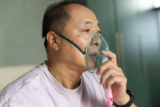 Senior Asian man holding oxygen nebulizer mask, wearing pink T-shirt in bedroom, gazing right