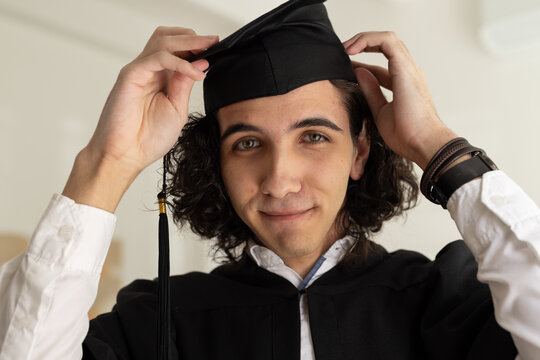 Male graduate adjusting mortarboard cap and tassel while standing in studio wearing gown, bracelets