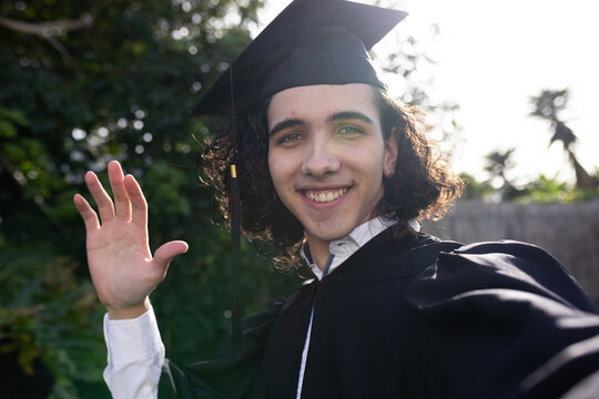 Male graduate waving and smiling chest-up in backyard with mortarboard tassel, gown and lens flare