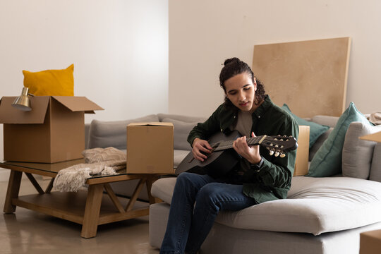 Adult male playing black acoustic guitar on gray sofa in living room with boxes, copy space