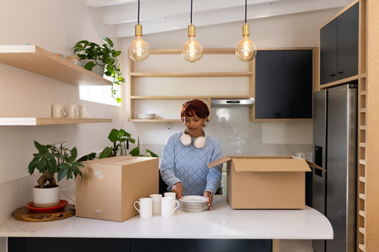 African American woman unpacking white plates and mugs from moving boxes in kitchen on countertop