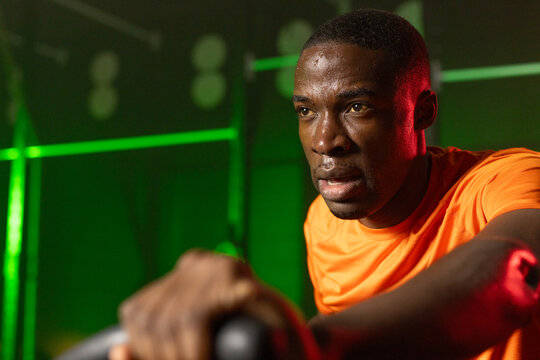 African adult man pushing spin bike leaning gripping bars in orange shirt under green-red lights