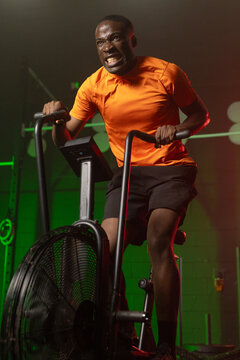 African American man pedaling hard on fan bike at gym, gripping upright handles in orange shirt