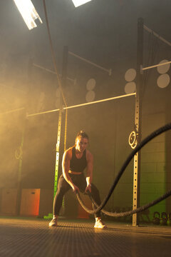 Female trainee gripping heavy battle ropes, swinging in low squat on rubber floor, wearing leggings