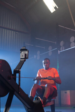 Adult man rowing on rowing machine in gym, wearing orange shirt and dark shorts, watching monitor