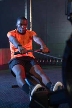 African American man rowing on ergometer at gym, pulling handle, wearing orange shirt, wristwatch