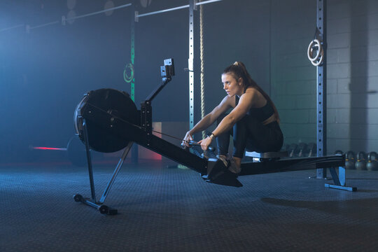 Female athlete in sportswear rowing on rower in gym, gripping handle, monitor visible, copy space
