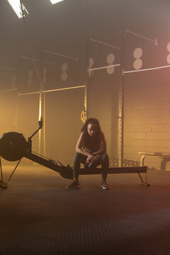 African American woman sitting on rower wearing sportswear holding water bottle resting in gym