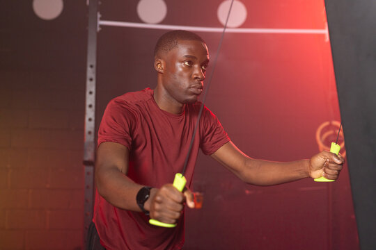 Adult African man performing cable pull gripping neon handles in gym wearing maroon tee smartwatch