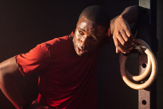 African man leaning against metal gym rig in gym, gripping wooden rings in red T-shirt