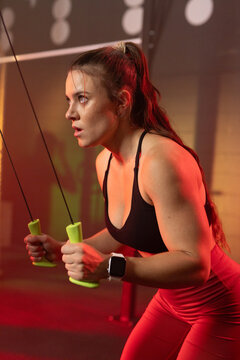 Female performing resistance exercise at gym pulling neon green cable handles wearing red leggings