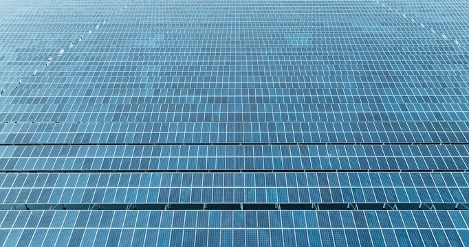 An aerial view shows rows of blue solar panels arranged in a dense grid pattern under bright daylight, highlighting sustainable energy.