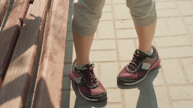 Person legs and feet in athletic shoes are shown walking on paved surface then resting by wooden bench. scene features consistent outdoor daylight with shadows visible underfoot and texture