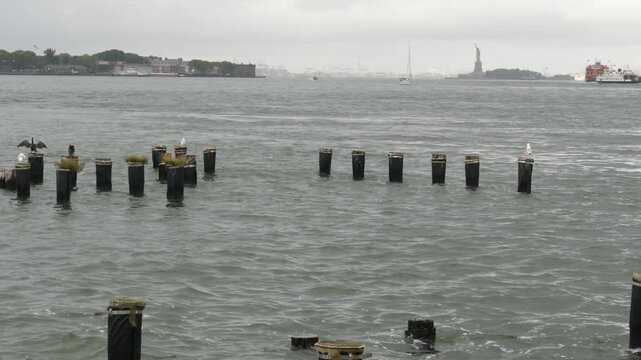 Statue of Liberty on Liberty Island from Brooklyn, New York City, United States. Ferry to Staten Island and birds on old wooden pier piles remains. Rainy foggy day weather. Orange boat water transport