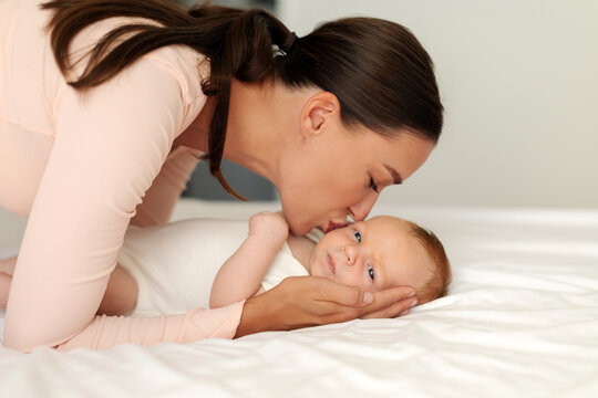 Mother lovingly kissing newborn baby lying on bed, tender side view portrait