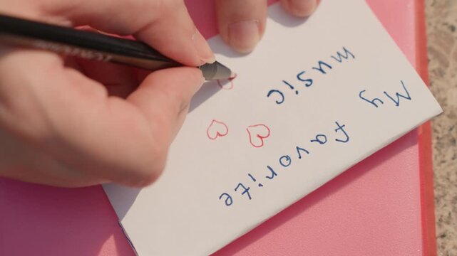 Hand writes on small white card with blue and red ink adding text like My favorite music alongside drawn hearts. scene is set against pink notebook or surface focusing closely on writing