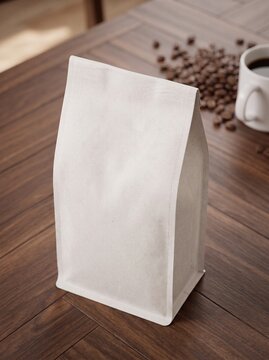 A plain white coffee bag stands upright on a dark wooden table, with scattered coffee beans and a white cup in the blurred background.