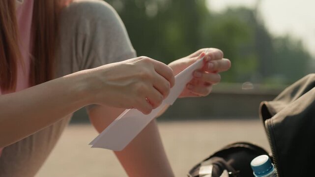 Young woman carefully folds piece of white paper likely making origami model. Woman sits outdoors with sunlight illuminating her hands and material against blurred green background