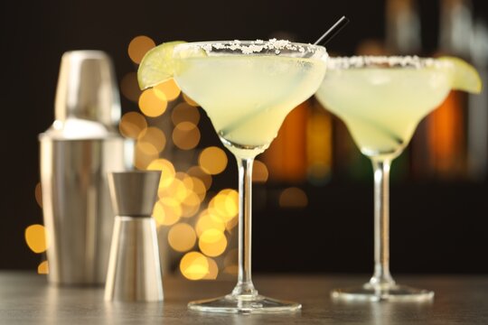 Delicious margarita cocktail in glasses, shaker and jigger on table against bokeh lights. Selective focus