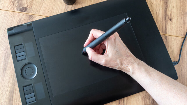 Hand holds a digital stylus and draws on a black graphics tablet. Creative process of creating a digital design