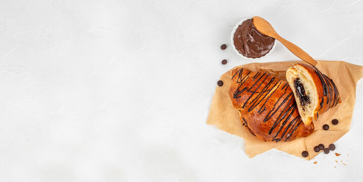 Chocolate croissant with drizzled chocolate on parchment paper, accompanied by a small bowl of chocolate spread and chocolate chips on a white background