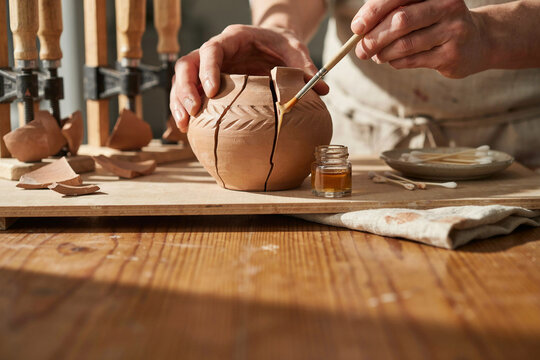 Hands repairing a cracked clay pot with glue and brush surrounded by broken shards