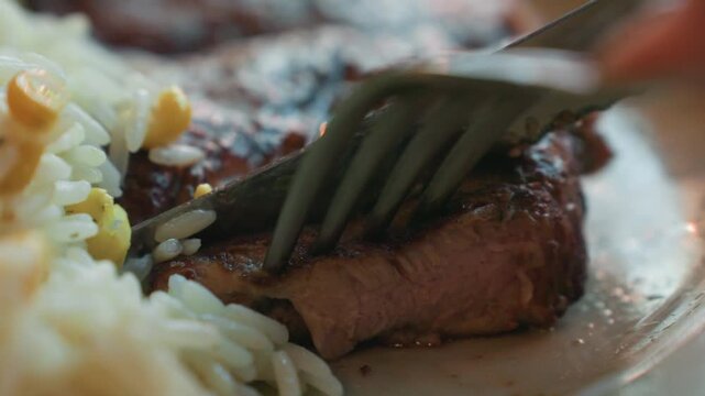 Close up view of cooked steak being cut with fork and knife accompanied by rice pilaf. Knife slices through meat while camera focuses on texture details like seared crust and juicy interior