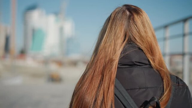 Woman with long brown hair walks away from camera through urban environment wearing black jacket and carrying bag. background features modern buildings under construction or renovation