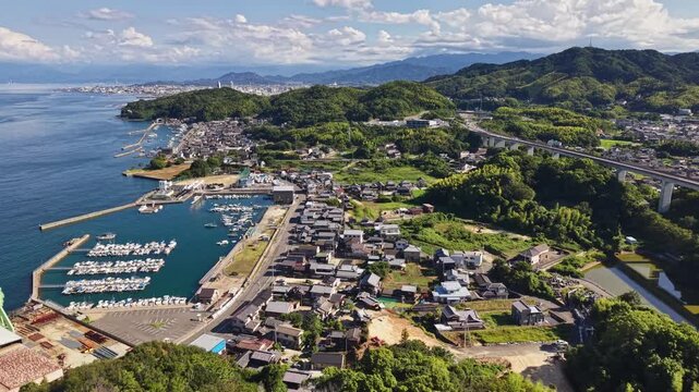 Aerial footage tracking over a scenic Japanese coastal village featuring a marina with docked boats, blue ocean water, and highway viaduct.
