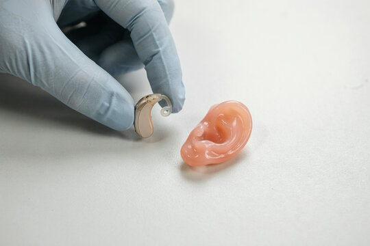 Gloved hand holding a behind the ear hearing aid beside a silicone ear model on white