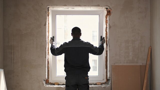 Professional installer fitting new window into renovated building opening with protective gloves securing white PVC frame during construction work polyurethane foam visible around perimeter rough bric