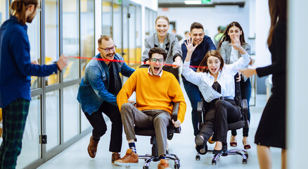 Fototapeta na wymiar Group of office colleagues cheerfully greets person sitting on chair as they are wheeled down hallway. Happy employees riding chairs in a bright corridor. Concept of office staff, workplace fun.